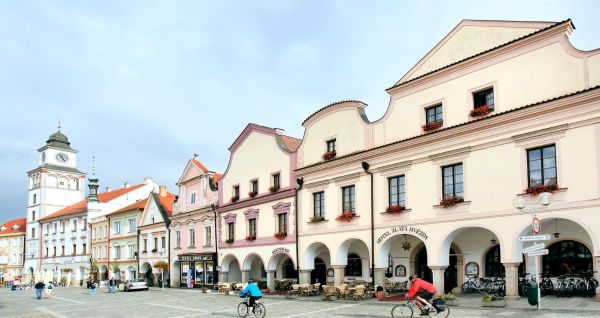 Zu sehen ist ein historischer Stadtplatz mit farbenfrohen Renaissancegebäuden, überdachten Arkaden und einladenden Sitzgelegenheiten im Freien. Unter bewölktem Himmel fahren zwei Menschen mit dem Fahrrad über die Kopfsteinpflasterstraße, während sich im H