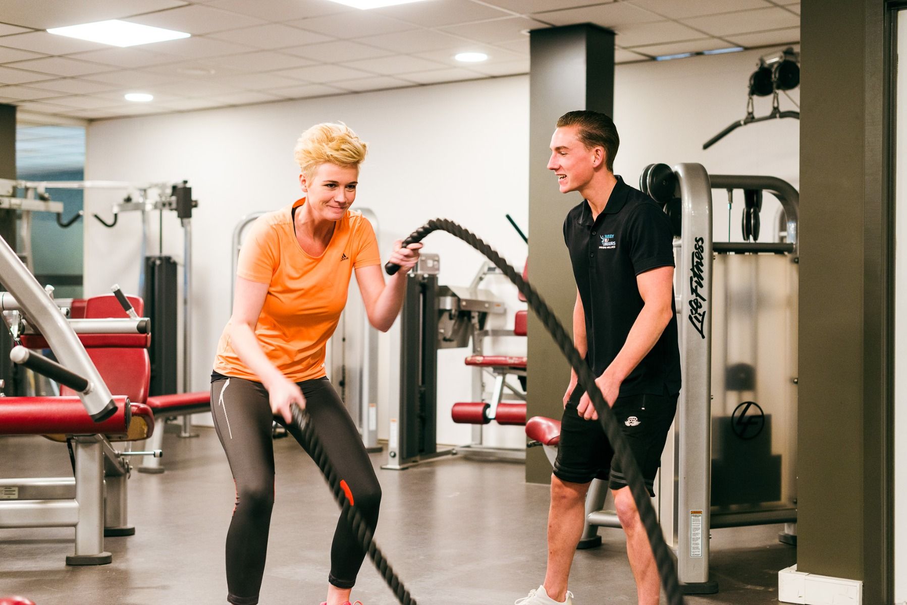 Eine Frau trainiert mit Kampfseilen im Sporthotel Bruurs, während ein Trainer in schwarzer Sportkleidung sie anspornt, während im Hintergrund verschiedene Fitnessgeräte zu sehen sind.
