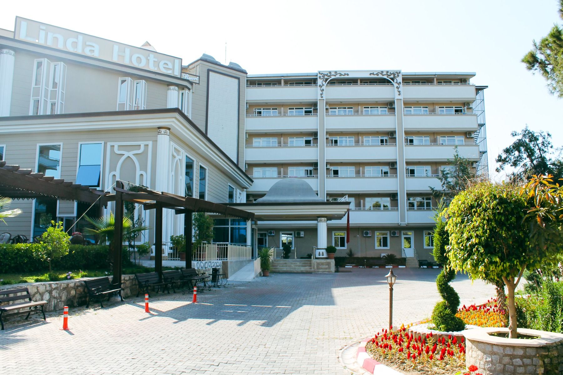Das mehrstöckige Hotel „Linda Resort“ verfügt über Balkone, einen mit Blumen und Bäumen begrünten Eingangsbereich und eine gepflasterte Zufahrt, die von orangefarbenen Verkehrskegeln gesäumt ist.
