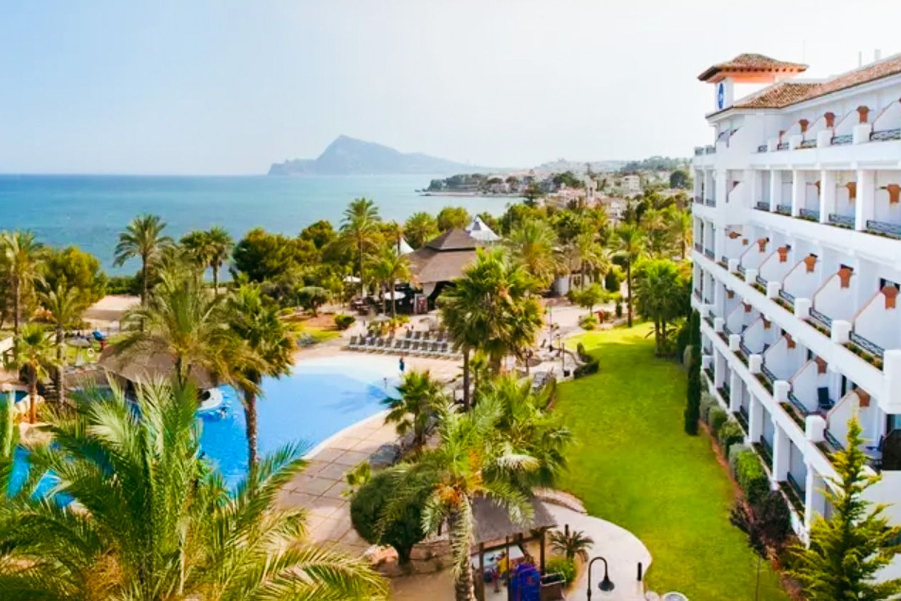 Das Hotel SH Villa Gadea* ist ein Resort an der Küste mit Meerblick, einem großen weißen Gebäude, einem Pool, Palmen und grünen Rasenflächen – ideal für Fußballmannschaften während ihres Trainingslagers in Altea unter strahlend blauem Himmel.