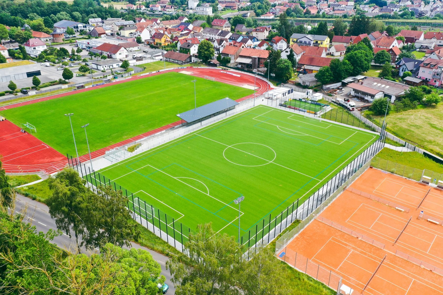 Luftaufnahme eines Sportkomplexes, der sich ideal für ein Trainingslager eignet, mit Fußballplatz, Laufbahn, Tennisplätzen und dem familiengeführten Landhotel Imhof in der Nähe, eingebettet zwischen Bäumen und Gebäuden in einer sonnigen Vorstadtlage.