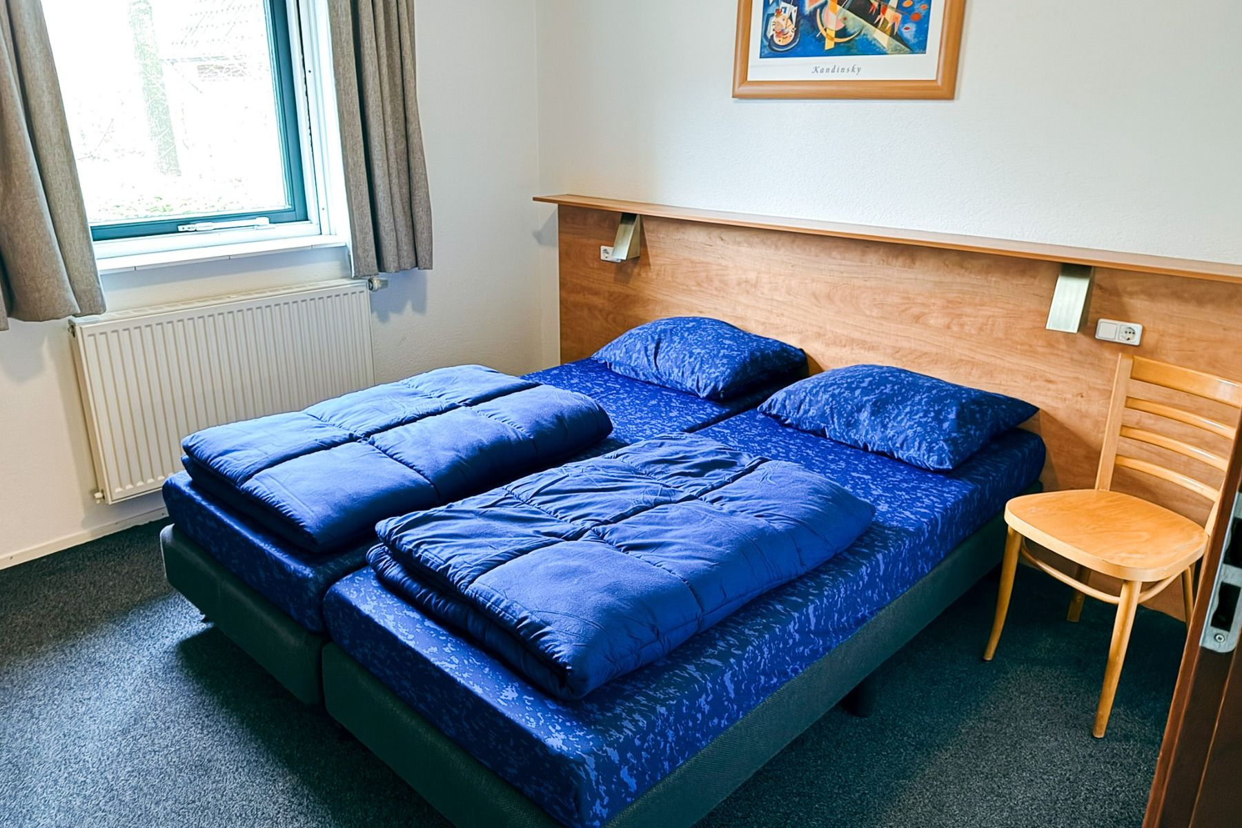 Ein Doppelbett mit blauem Bettzeug steht an einem hölzernen Kopfteil in einem hellen, kompakten Zimmer in Marveld Recreatie. In der Nähe steht ein Holzstuhl mit einem Fenster und Vorhängen über dem Heizkörper, die an den gemütlichen Charme der Ferienpark-