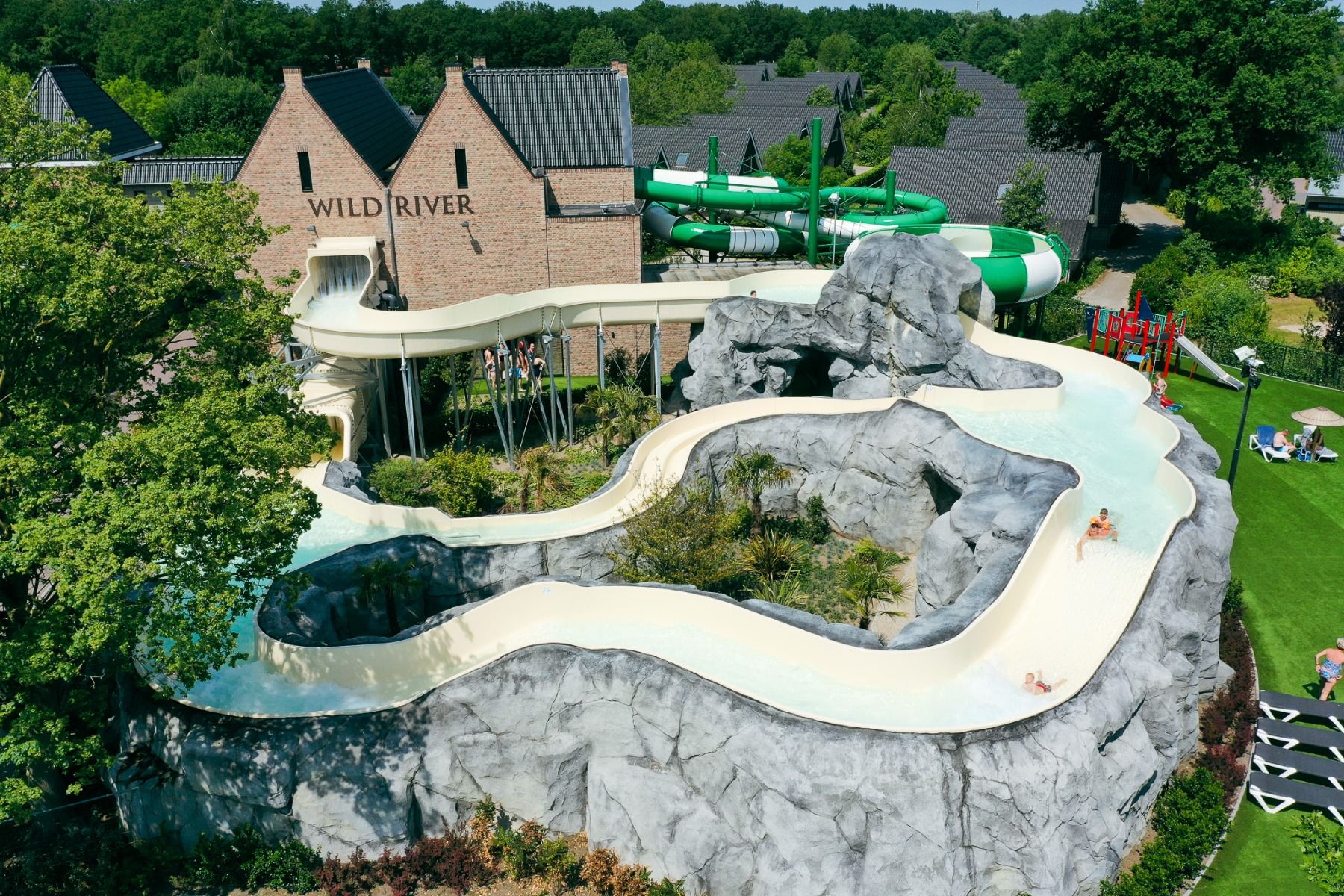 Eine kurvenreiche Wasserrutsche namens Wild River schlängelt sich um künstliche Felsen im Marveld Recreatie, umgeben von Grünflächen, Bungalows und Gebäuden. Mehrere Personen rutschen hinunter, während im Hintergrund grüne Röhrenrutschen zu sehen sind.
