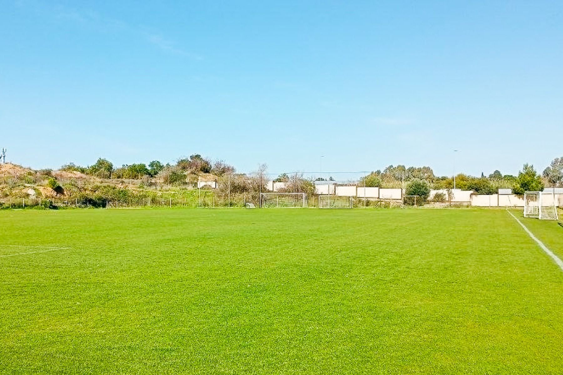 Ein strahlender, sonniger Tag auf dem leeren Fußballplatz des Side Moon Palace, mit grünem Rasen, weißen Torpfosten und einer Kulisse aus Bäumen und Sträuchern unter einem strahlend blauen Himmel.