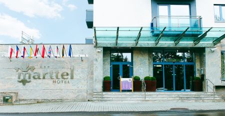 Das Hotel Marttel*** ist ein modernes wei&szlig;es Geb&auml;ude mit vielen Fenstern in der N&auml;he des gr&uuml;nen Kunstrasen-Fu&szlig;ballplatzes, umgeben von B&auml;umen und H&uuml;geln, mit Wohnh&auml;usern im Hintergrund unter einem teilweise bew&ouml;lkten Himmel, nahe der deutschen Grenze.