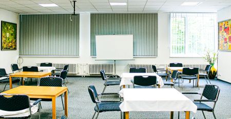 Das Hotel Marttel*** ist ein modernes wei&szlig;es Geb&auml;ude mit vielen Fenstern in der N&auml;he des gr&uuml;nen Kunstrasen-Fu&szlig;ballplatzes, umgeben von B&auml;umen und H&uuml;geln, mit Wohnh&auml;usern im Hintergrund unter einem teilweise bew&ouml;lkten Himmel, nahe der deutschen Grenze.