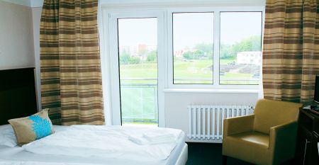 Das Hotel Marttel*** ist ein modernes wei&szlig;es Geb&auml;ude mit vielen Fenstern in der N&auml;he des gr&uuml;nen Kunstrasen-Fu&szlig;ballplatzes, umgeben von B&auml;umen und H&uuml;geln, mit Wohnh&auml;usern im Hintergrund unter einem teilweise bew&ouml;lkten Himmel, nahe der deutschen Grenze.