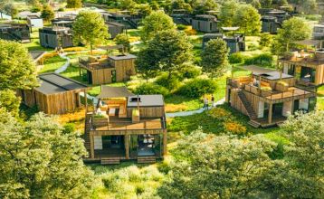 Im Resort De Parel öffnen sich moderne Holzkabinen mit großen Glastüren zu einem Deck mit Stühlen, einem Tisch und einem weißen Sonnenschirm - perfekt für Teambuilding oder Trainingslager im Grünen und ähnliche Kabinen unter teilweise bewölktem Himmel.