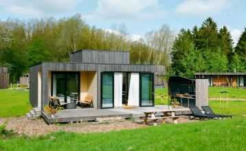 Im Resort De Parel öffnen sich moderne Holzkabinen mit großen Glastüren zu einem Deck mit Stühlen, einem Tisch und einem weißen Sonnenschirm - perfekt für Teambuilding oder Trainingslager im Grünen und ähnliche Kabinen unter teilweise bewölktem Himmel.