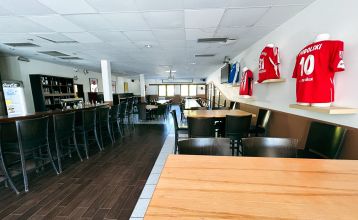 Ein heller Raum im Sportpark Fun4You ist mit runden und rechteckigen Tischen, schwarzen Stühlen, großen Fenstern, einem an der Wand montierten TV-Projektor und einem "Bundesliga"-Schild ausgestattet. Mit Blick auf den Sportplatz ist er perfekt für Teambui