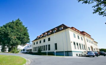 Das Gästehaus Burgenland Güssing ist ein großes, cremefarbenes Gebäude mit rotem Dach und vielen Fenstern unter einem strahlend blauen Himmel, vor dem mehrere Autos geparkt sind und in dessen Nähe Bäume stehen.