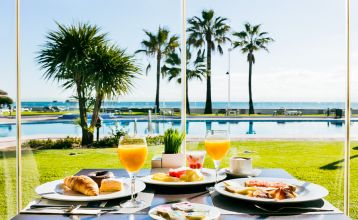 Ein hohes, gelbes Hotel Sol Guadalmar mit Balkonen blickt auf einen klaren, blauen Außenpool, umgeben von Palmen und grünen Rasenflächen unter einem sonnigen Himmel - perfekt für Ihren Aufenthalt in diesem zentrumsnahen 4 Sterne Hotel Malaga.