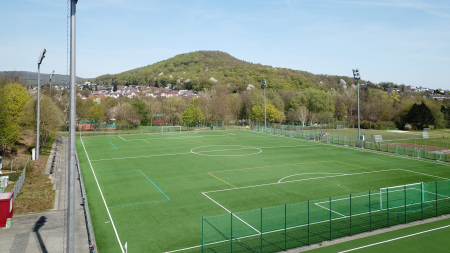 Der Kunstrasenplatz Baunatal ist ein weitläufiger, künstlich angelegter und eingezäunter Fußballplatz vor der Kulisse von Bäumen, Häusern und einem baumbestandenen Hügel unter einem strahlend blauen Himmel.