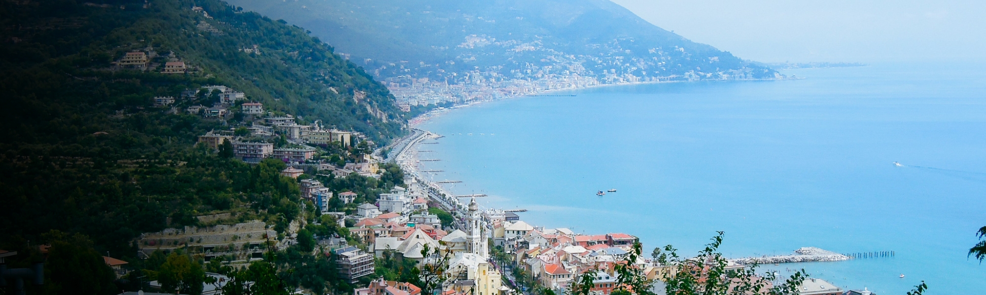 Panoramaansicht der Italienischen Riviera mit der Küstenstadt, die sich entlang des blauen Meeres erstreckt, umgeben von hügeliger Landschaft und grüner Vegetation.