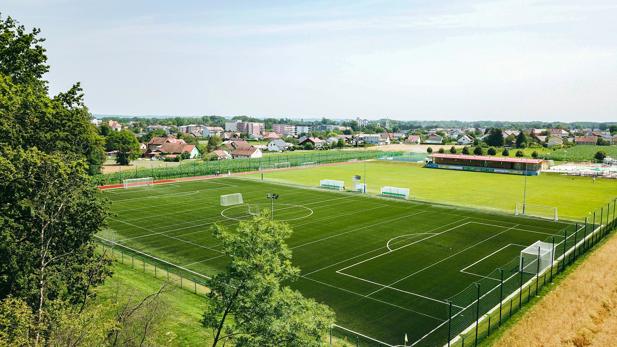 Fußballtrainingslager mit Kunstrasen- und Naturrasenplatz inmitten einer idyllischen Landschaft mit kleinen Häusern im Hintergrund.