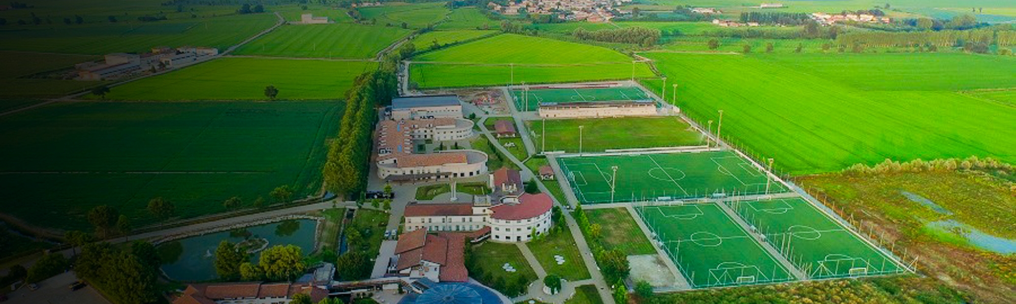 Luftaufnahme einer weitläufigen Sportanlage im Piemont, umgeben von grünen Feldern, mit mehreren Fußballplätzen, Gebäuden und Trainingsmöglichkeiten.