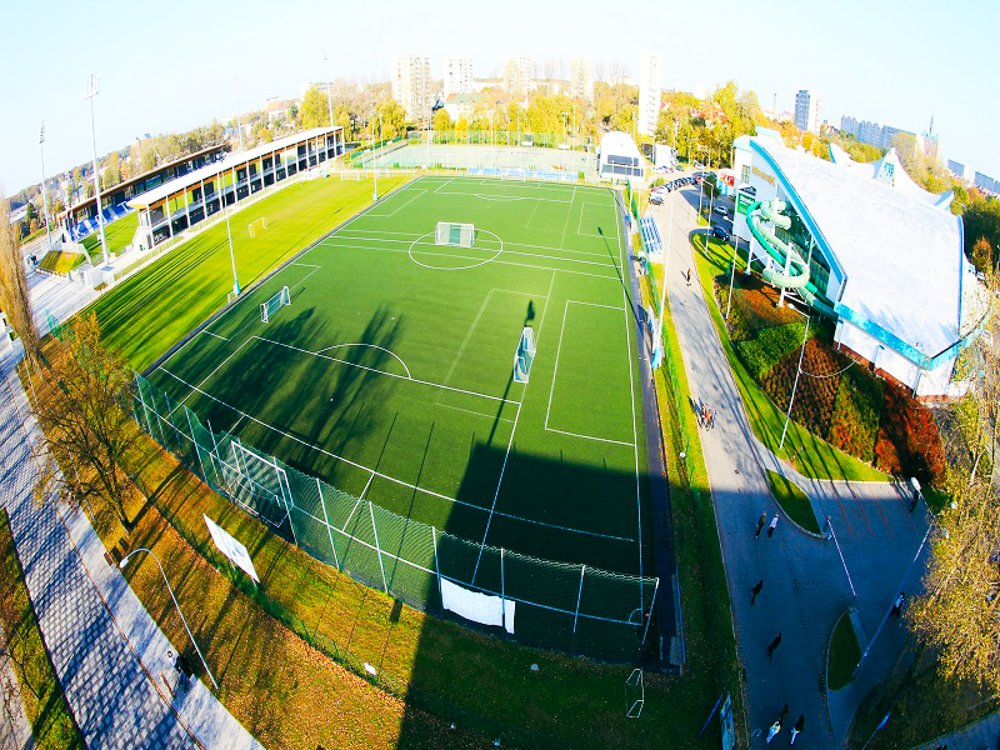 Luftaufnahme einer modernen Fußballtrainingsanlage in Polen mit Kunstrasenplatz, Tribüne und angrenzendem Freizeitbad.