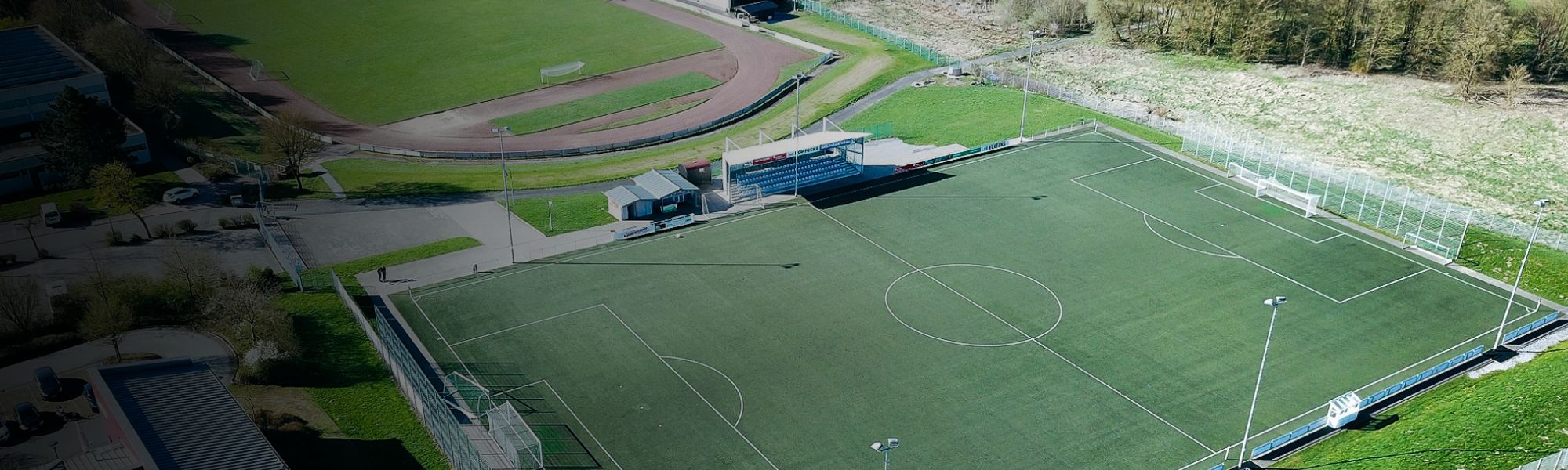 Kunstrasen-Fußballplatz auf Sardinien mit markierten Spielfeldlinien und Umzäunung, im Hintergrund Berge und Gebäude.