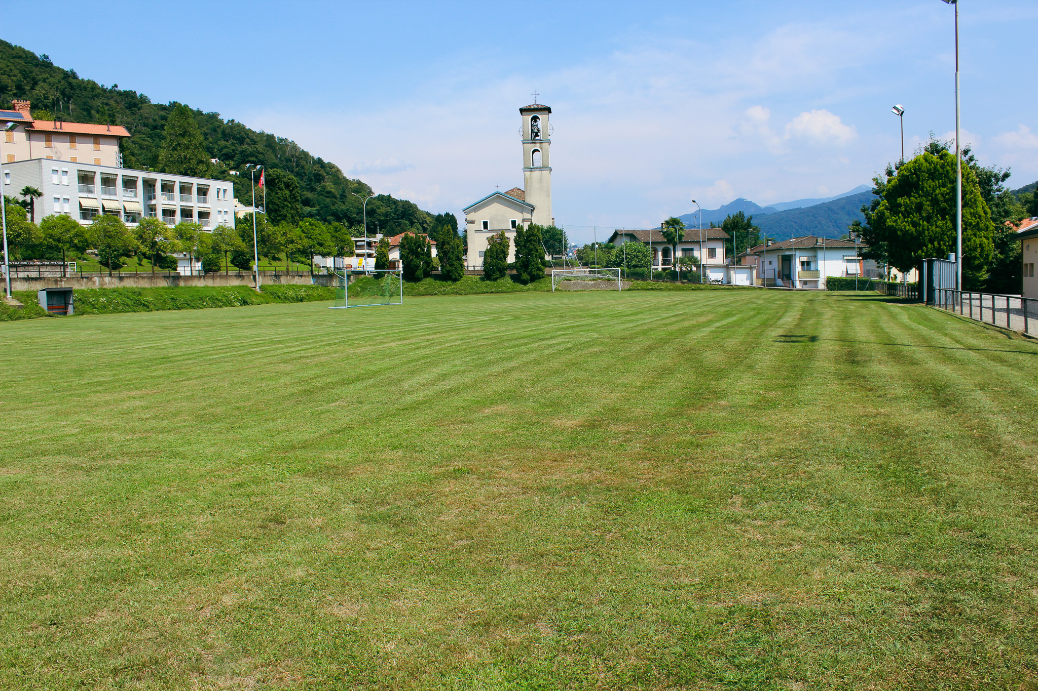 Ein gepflegter Fußballplatz in der malerischen Region Tessin, umgeben von grünen Hügeln und traditioneller Architektur. Im Hintergrund eine Kirche und Wohngebäude, die eine idyllische Kulisse für ein Fußballtrainingslager bieten.