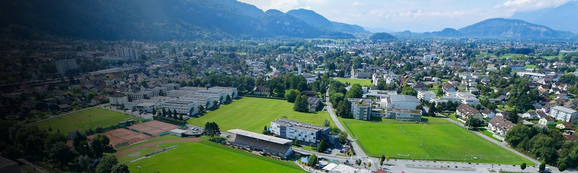 Panoramaaufnahme eines Sportgeländes in Vorarlberg mit Fußballplätzen, Stadiontribüne und angrenzenden Gebäuden, eingebettet in eine bergige Landschaft.