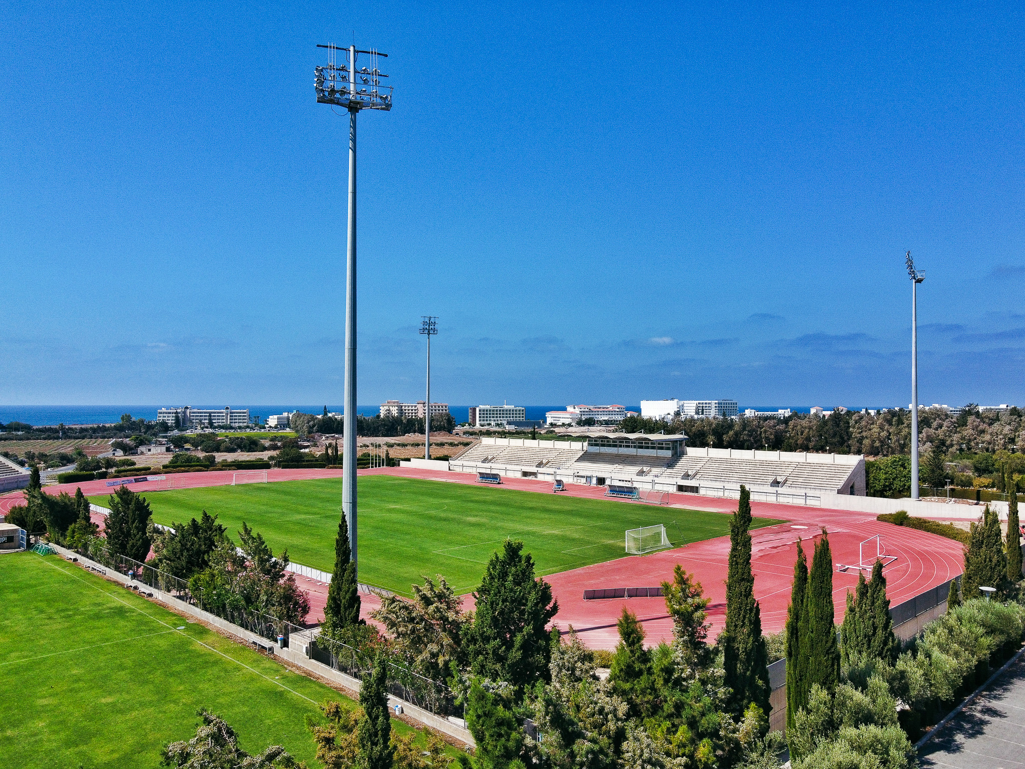 Sportstadion auf Zypern mit Flutlichtmasten, Laufbahn und Fußballfeld, mit Aussicht auf das Mittelmeer und moderne Hotelbauten.