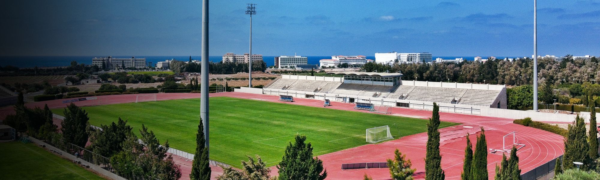 Fußballplatz mit Laufbahn und Tribüne auf Zypern, umgeben von Bäumen, mit Blick auf das Meer und Hotelanlagen im Hintergrund.