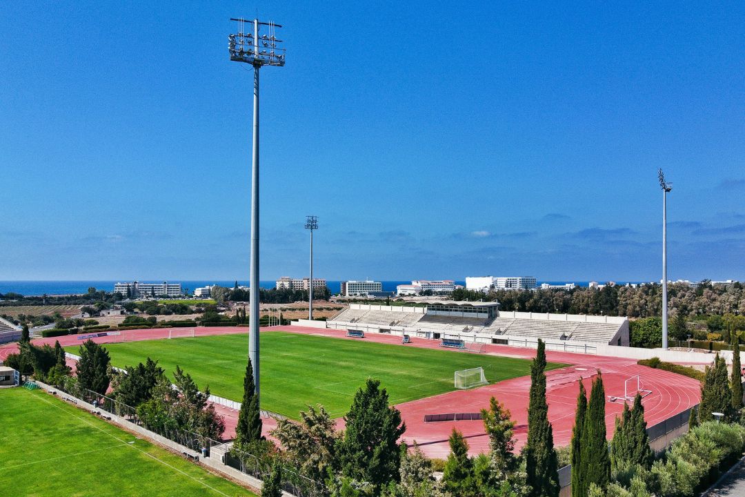 Luftaufnahme eines modernen Fußballstadions auf Zypern mit Laufbahn, Tribünen und Meerblick im Hintergrund.