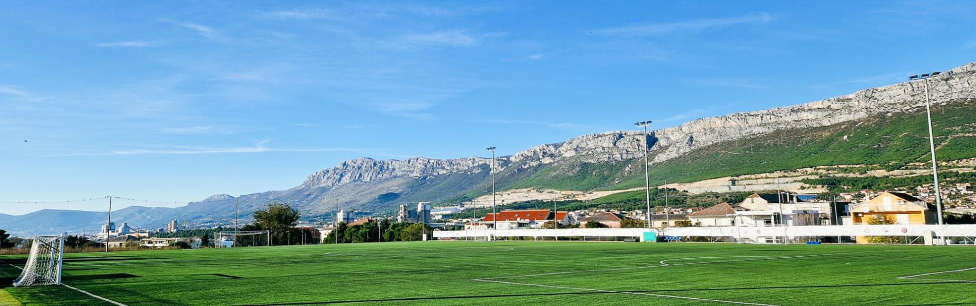 Sportanlage in Dalmatien mit einem Fu&szlig;ballplatz, Flutlichtmasten und Blick auf eine malerische Bergkette sowie Wohnh&auml;user im Hintergrund.
