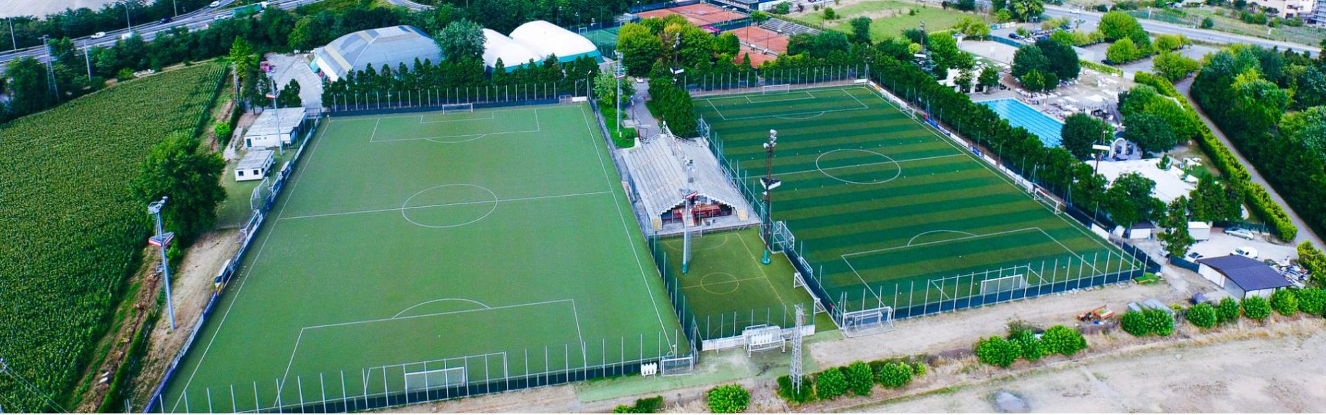 Luftaufnahme einer Sportanlage in der Lombardei mit mehreren Fu&szlig;ballpl&auml;tzen, Tennispl&auml;tzen, einer Trib&uuml;ne und einer Schwimmhalle, umgeben von landwirtschaftlichen Feldern und einer kleinen Ortschaft.