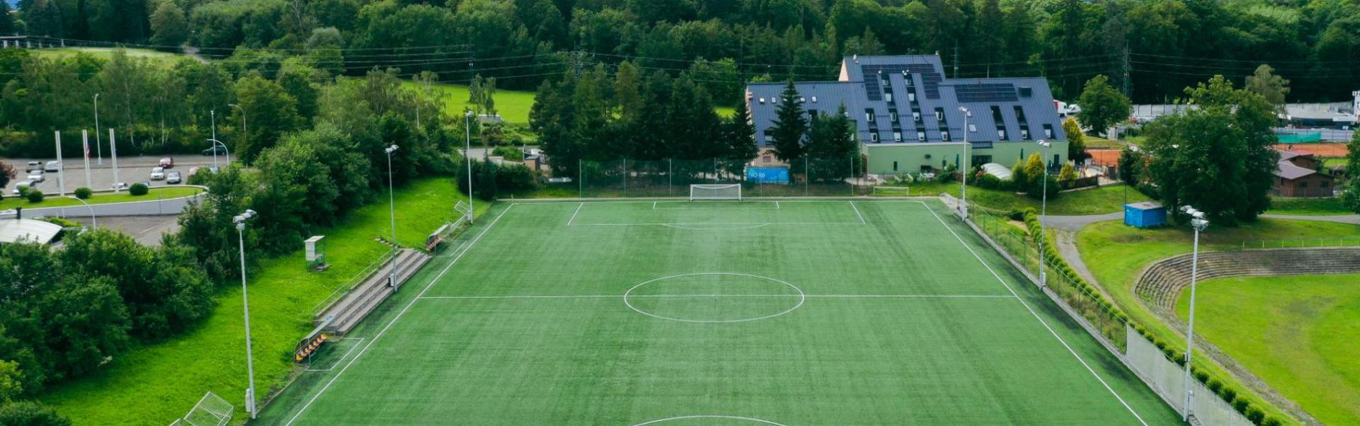 Fu&szlig;ballplatz mit Kunstrasen in einer idyllischen Umgebung, angrenzend an ein modernes Geb&auml;ude.