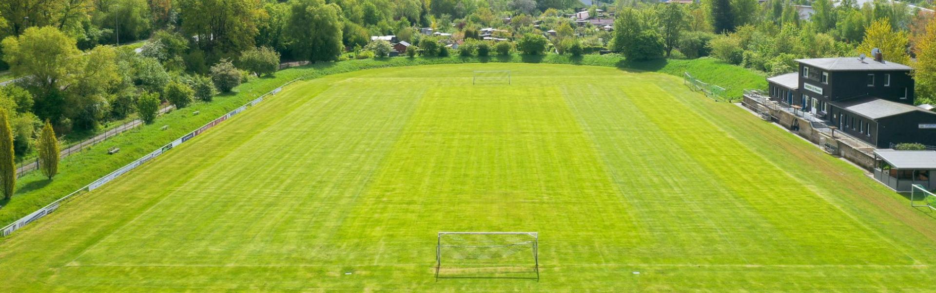 Luftaufnahme eines Fu&szlig;ballplatzes mit gepflegtem Rasen in Niedersachsen, umgeben von viel Gr&uuml;n, mit einem Vereinsheim auf der rechten Seite.