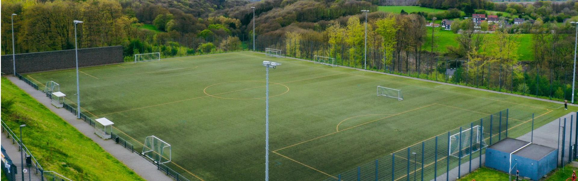  Weitl&auml;ufiges Fu&szlig;balltrainingsgel&auml;nde mit Kunstrasenplatz in Nordrhein-Westfalen, eingebettet in eine malerische Naturlandschaft mit dichten W&auml;ldern und H&uuml;geln.