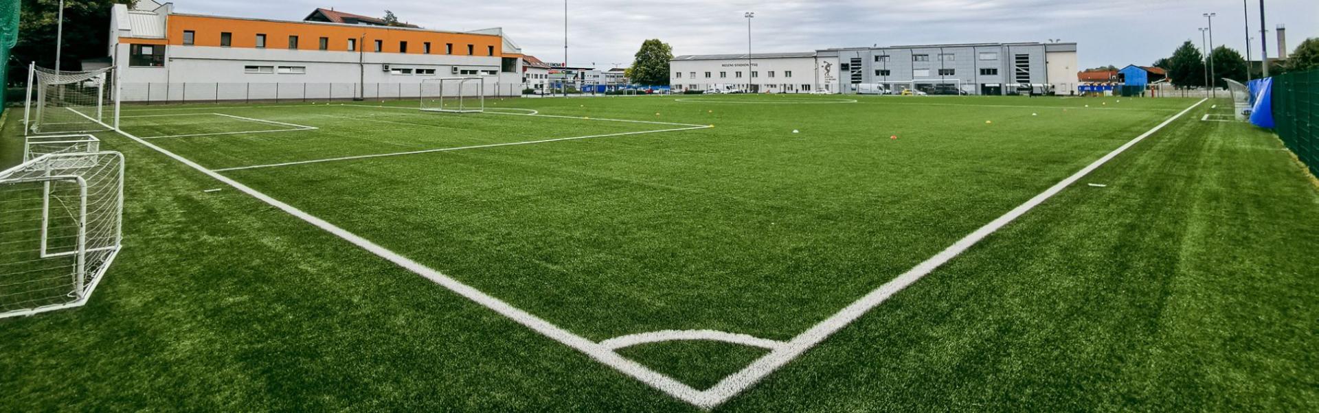 Kunstrasen-Fußballplatz mit Flutlichtanlage umgeben von Häusern.
