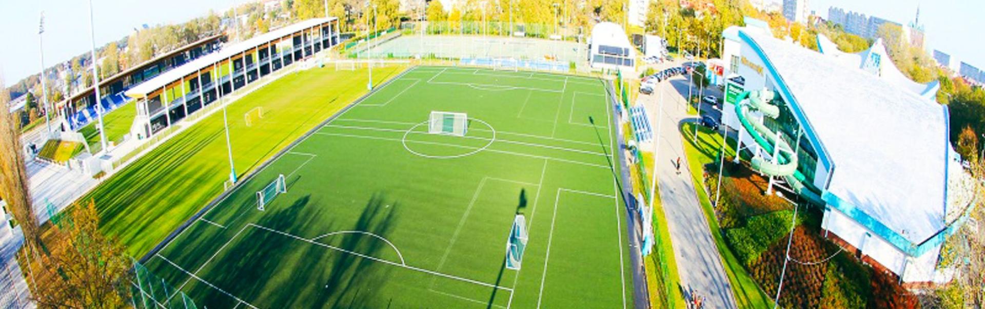 Luftaufnahme eines Sportkomplexes im Freien mit einem gr&uuml;nen Kunstrasen-Fu&szlig;ballfeld, Zuschauertrib&uuml;nen auf der linken Seite und einem modernen Geb&auml;ude mit einer gro&szlig;en Drachenskulptur auf der rechten Seite. Im Hintergrund sind B&auml;ume und st&auml;dtische Geb&auml;ude