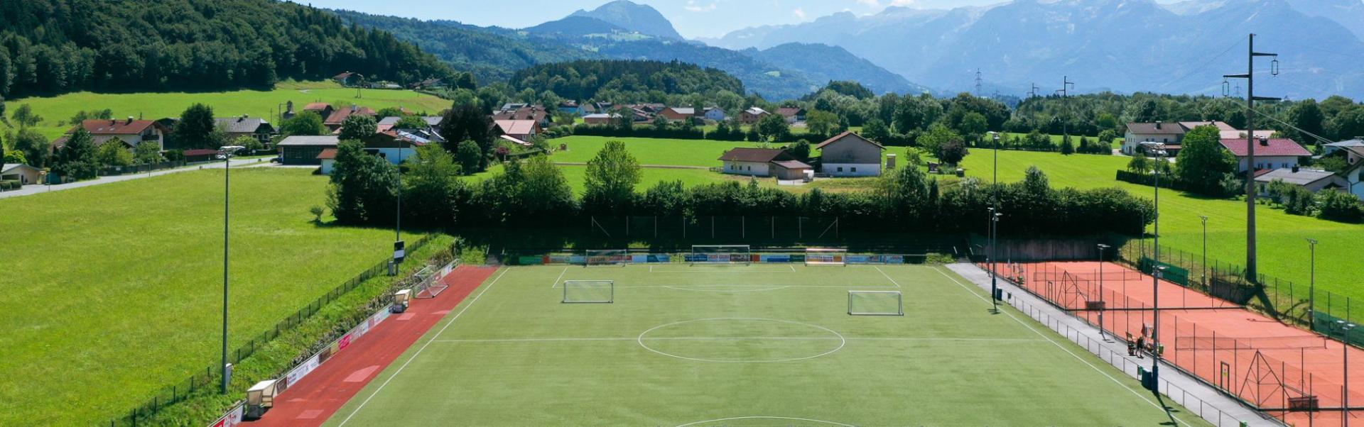 Fu&szlig;balltrainingsanlage in Salzburg mit Kunstrasenplatz, roten Laufbahnen und Tennispl&auml;tzen, eingebettet in eine idyllische Alpenlandschaft.