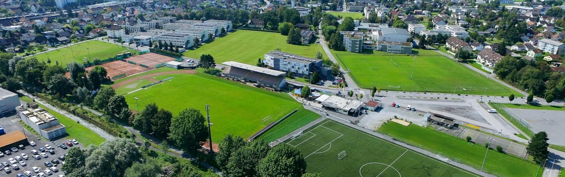 Panoramaaufnahme eines Sportgel&auml;ndes in Vorarlberg mit Fu&szlig;ballpl&auml;tzen, Stadiontrib&uuml;ne und angrenzenden Geb&auml;uden, eingebettet in eine bergige Landschaft.