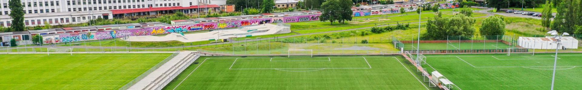  Fu&szlig;ballplatz mit Kunstrasen in einer gr&uuml;nen Umgebung, umgeben von H&uuml;geln und einer Stadtlandschaft im Hintergrund.