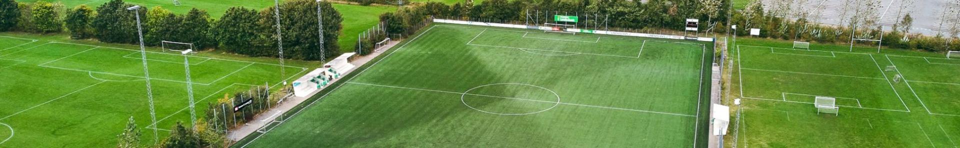 Luftaufnahme einer modernen Fu&szlig;balltrainingsanlage in D&auml;nemark mit einem gro&szlig;en Kunstrasenplatz, mehreren Naturrasenpl&auml;tzen, Flutlichtanlagen und angrenzenden Industrie- und Gewerbegeb&auml;uden im Hintergrund.