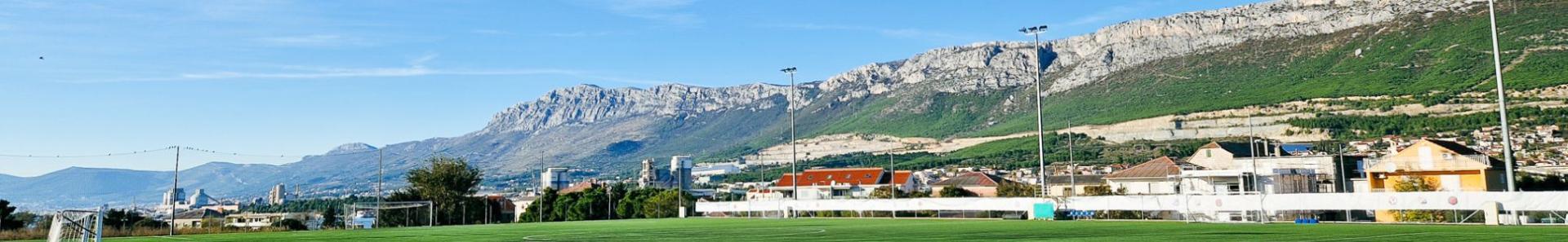 Sportanlage in Dalmatien mit einem Fu&szlig;ballplatz, Flutlichtmasten und Blick auf eine malerische Bergkette sowie Wohnh&auml;user im Hintergrund.