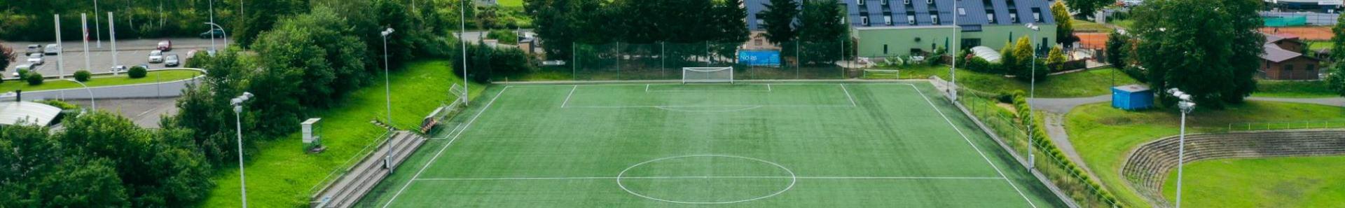 Fu&szlig;ballplatz mit Kunstrasen in einer idyllischen Umgebung, angrenzend an ein modernes Geb&auml;ude.