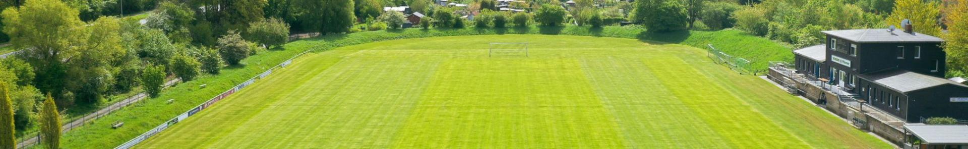 Fu&szlig;ballplatz in Niedersachsen mit frisch gem&auml;htem Rasen, eingerahmt von B&auml;umen einem modernen Vereinsheim mit Terrasse.