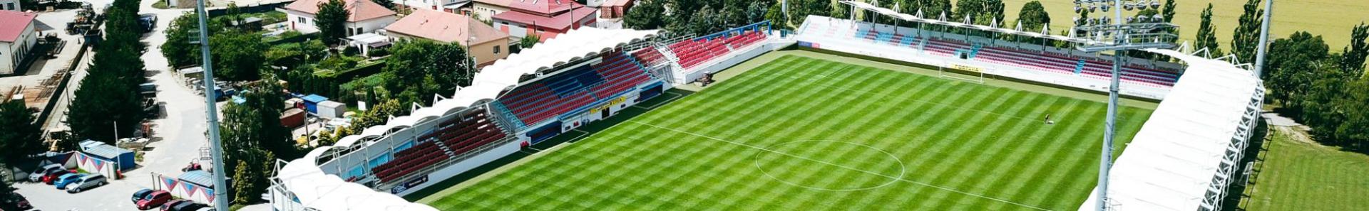 Fu&szlig;ballstadion in l&auml;ndlicher Umgebung mit Trib&uuml;nen, Flutlichtanlage und einer weiten Landschaft im Hintergrund.