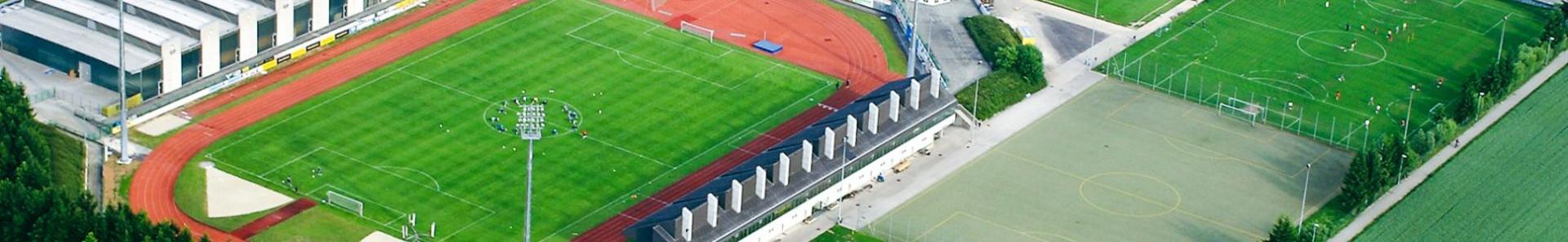 Luftaufnahme einer modernen Sportanlage in Ober&ouml;sterreich mit mehreren Fu&szlig;ballpl&auml;tzen, darunter ein Stadion mit Laufbahn, Kunstrasen- und Rasenpl&auml;tzen, umgeben von Parkpl&auml;tzen, Gewerbebauten und Wohnh&auml;usern.