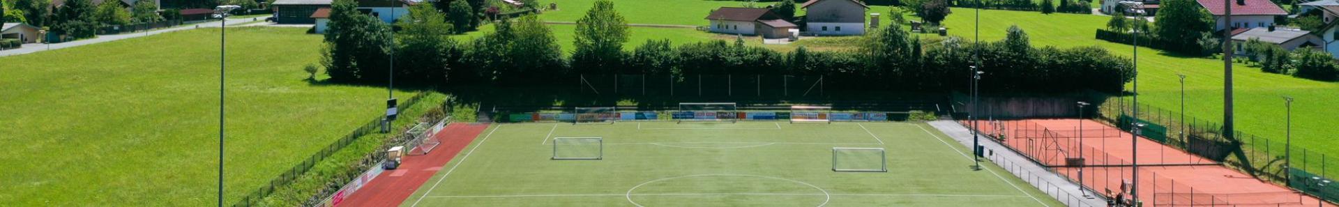 Fu&szlig;ballplatz in Salzburg, umgeben von gr&uuml;nen Wiesen, kleinen H&auml;usern und malerischen Bergen im Hintergrund, unter blauem Himmel.