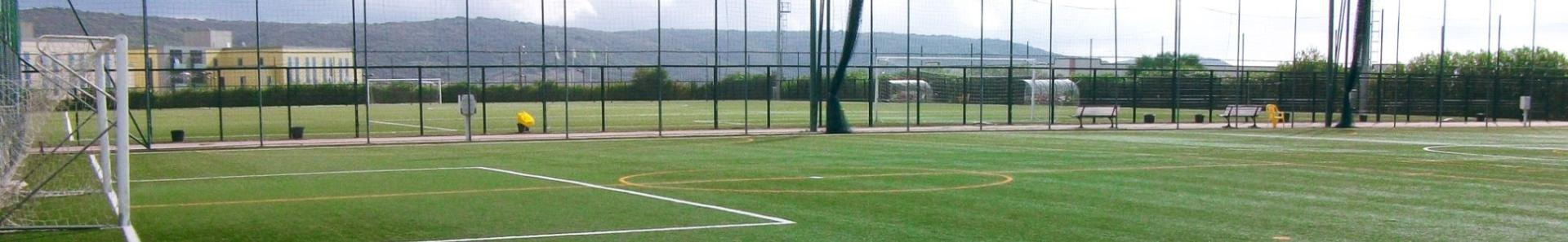 Fu&szlig;balltrainingsplatz auf Sardinien mit gr&uuml;nem Kunstrasen, Tor, B&auml;nken und Flutlichtmasten, umgeben von Netzen und Berglandschaft.
