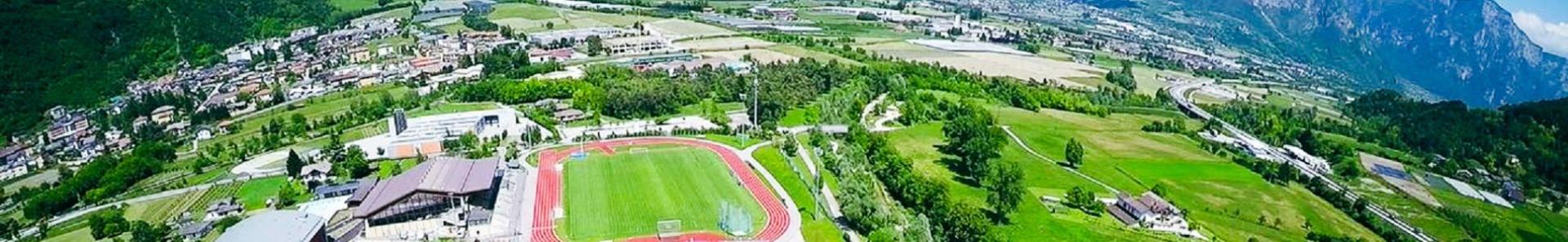 Sportanlage in Trentino mit Fu&szlig;ballplatz, Leichtathletikstadion, Sporthalle und gr&uuml;nen Feldern, eingebettet in eine idyllische Berglandschaft.