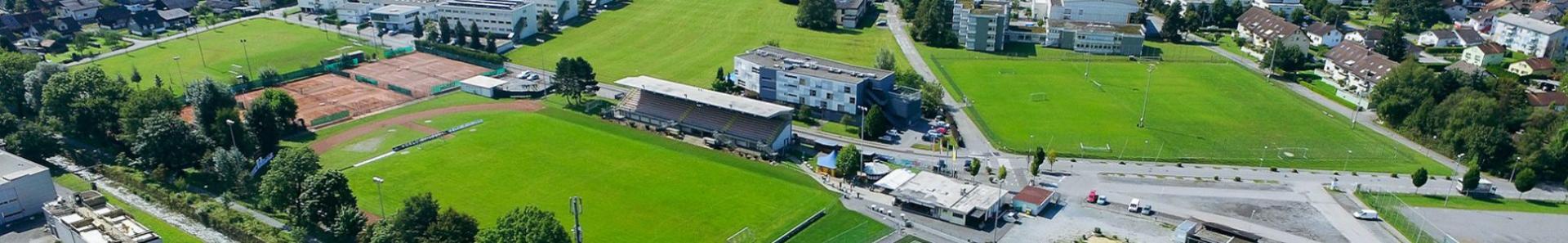 Luftaufnahme einer Sportanlage in Vorarlberg, umgeben von einer Stadt und Bergen im Hintergrund, mit mehreren Fu&szlig;ballpl&auml;tzen, Tennispl&auml;tzen und Sporteinrichtungen.