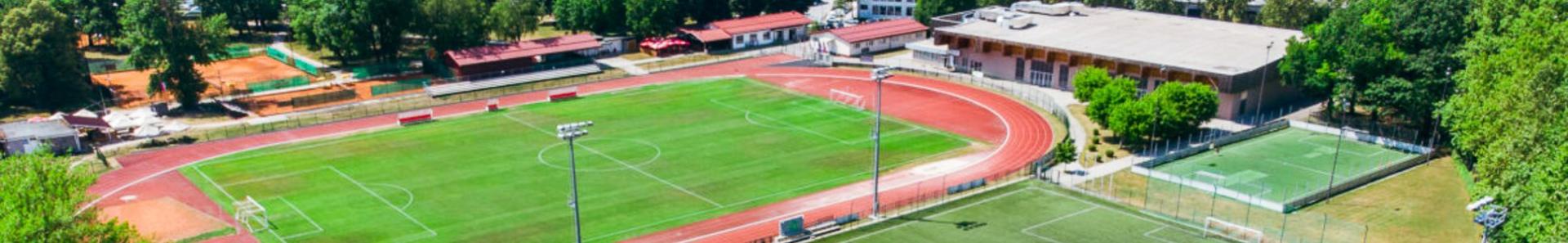 Fu&szlig;balltrainingslager mit mehreren Spielfeldern, Laufbahn und Tennispl&auml;tzen, eingebettet in eine st&auml;dtische Umgebung mit Bergen im Hintergrund.