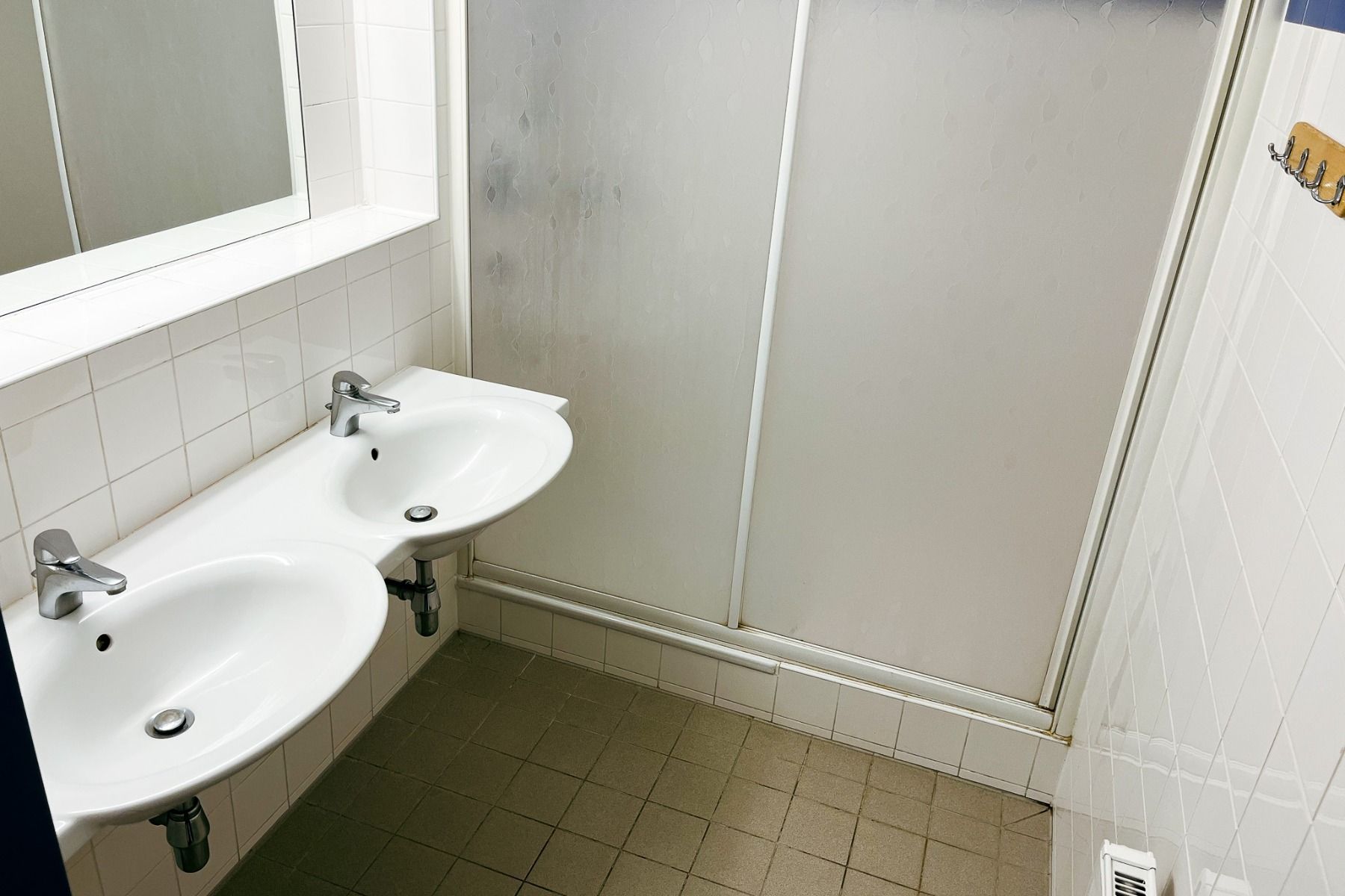 Das Gästehaus Burgenland Güssing verfügt über ein sauberes Badezimmer mit zwei weißen Waschbecken, einem Wandspiegel, gefliesten Wänden und Böden, einer Duschtür aus Milchglas und einem Kleiderständer aus Holz.