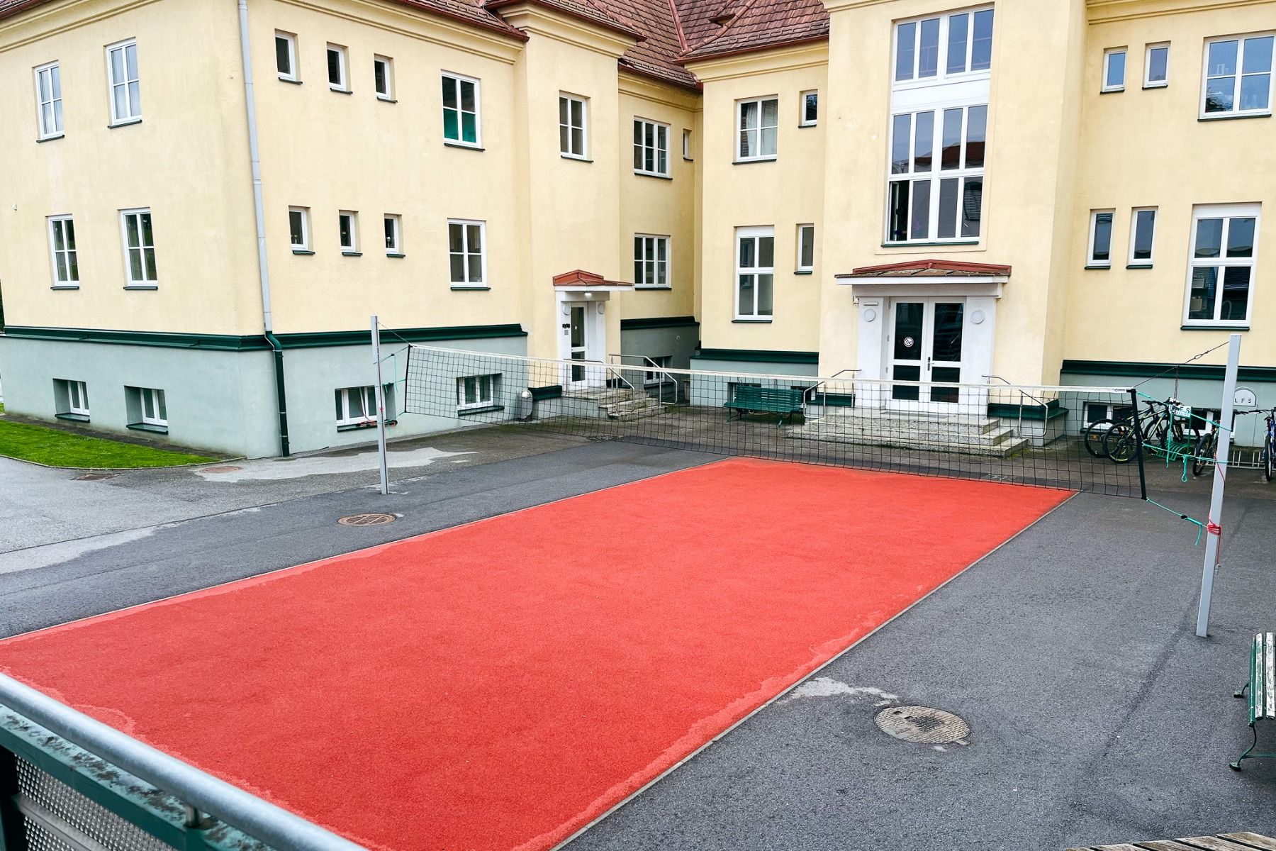 Vor dem Gästehaus Burgenland Güssing, einem gelben Gebäude mit mehreren Fenstern, zwei weißen Eingangstüren und rechts geparkten Fahrrädern, ist ein kleiner Volleyballplatz mit rotem Belag und Netz aufgebaut.